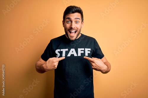 Young handsome worker man with beard wearing staff uniform t-shirt over yellow background very happy pointing with hand and finger