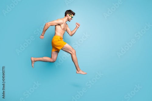 Full length body size profile side view of nice attractive cheerful purposeful guy in swimming shorts jumping running fast isolated on bright vivid shine vibrant green blue turquoise color background