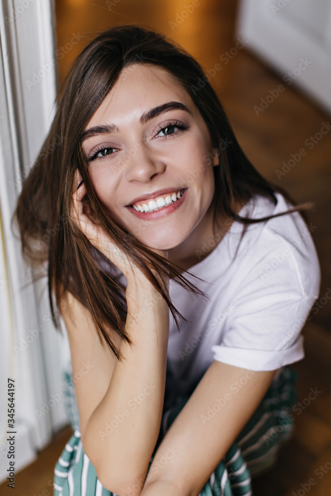 Cute dark-eyed girl smiling to camera while chilling at home. Indoor overhead photo of brunette attractive woman in white t-shirt. Cute dark-eyed girl smiling to camera while chilling at home. Indoor overhead photo of brunette attractive woman in white t-shirt.