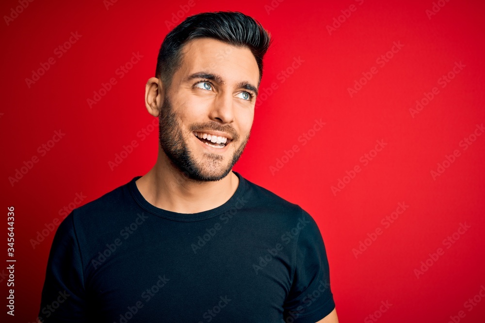 Young handsome man wearing casual black t-shirt standing over isolated red background looking away to side with smile on face, natural expression. Laughing confident.