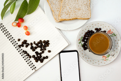 Top view of coffee beans and carissa carandas on notebook,leaves,two pieces of bread,mobile phone,and cup of coffee with saucer,isolated on background.