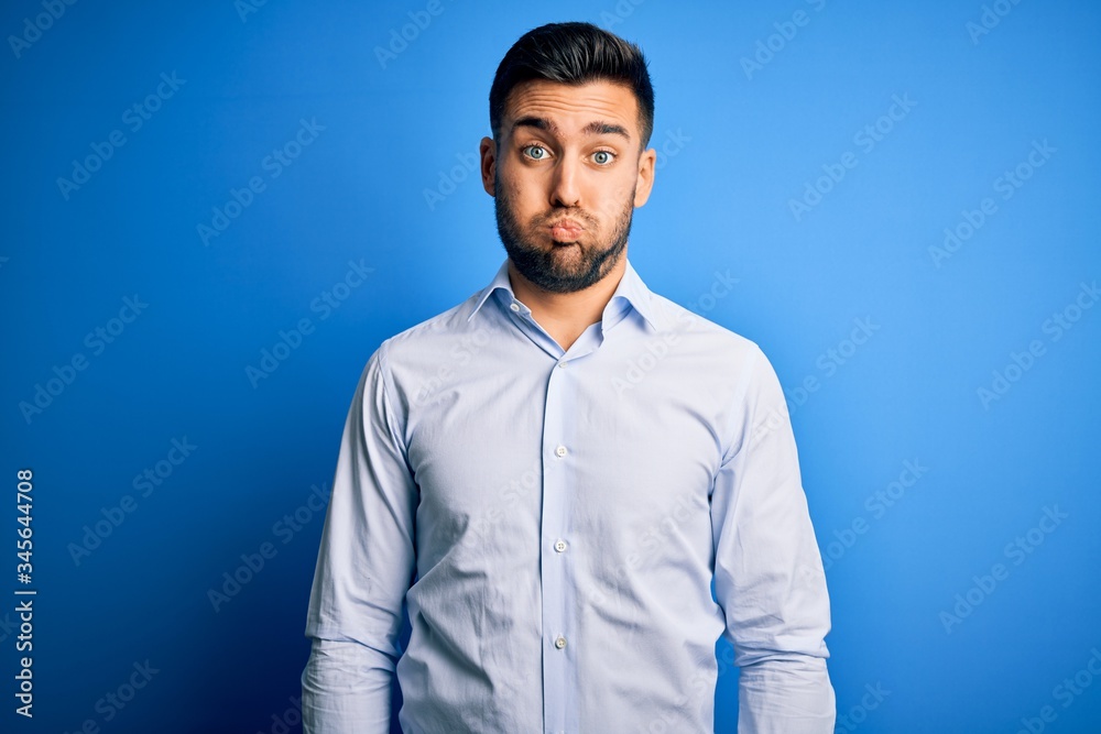 Young handsome man wearing elegant shirt standing over isolated blue background puffing cheeks with funny face. Mouth inflated with air, crazy expression. Young handsome man wearing elegant shirt standing over isolated blue background puffing cheeks with funny face. Mouth inflated with air, crazy expression.
