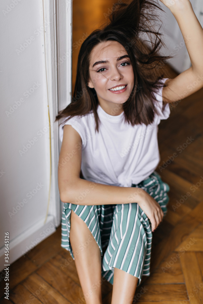 Tanned fascinating female model sitting on the floor and playing with her shiny hair. Indoor photo of lovable girl relaxing at home. Tanned fascinating female model sitting on the floor and playing with her shiny hair. Indoor photo of lovable girl relaxing at home.