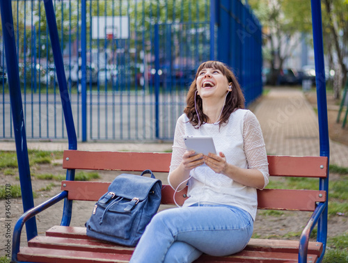 The girl is sitting on a bench with a tablet and laughing  looking up. Brunette girl in a white jacket and jeans on the street