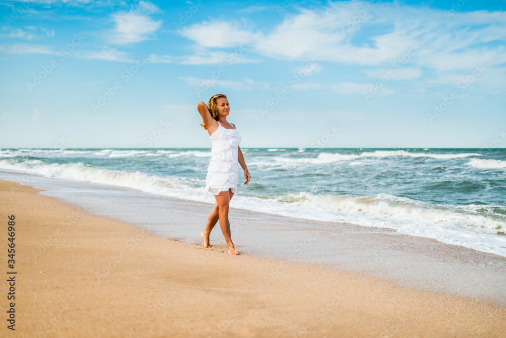 Charming young woman in a white dress walks along the calm sea waves on the sandy coast against a background of blue sky. White clouds on a sunny warm summer day