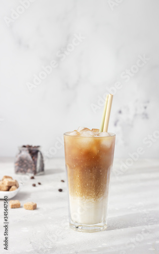 Cold coffee with milk and ice cubes in a tall glass with bamboo straw, light grey background. Selective focus. 