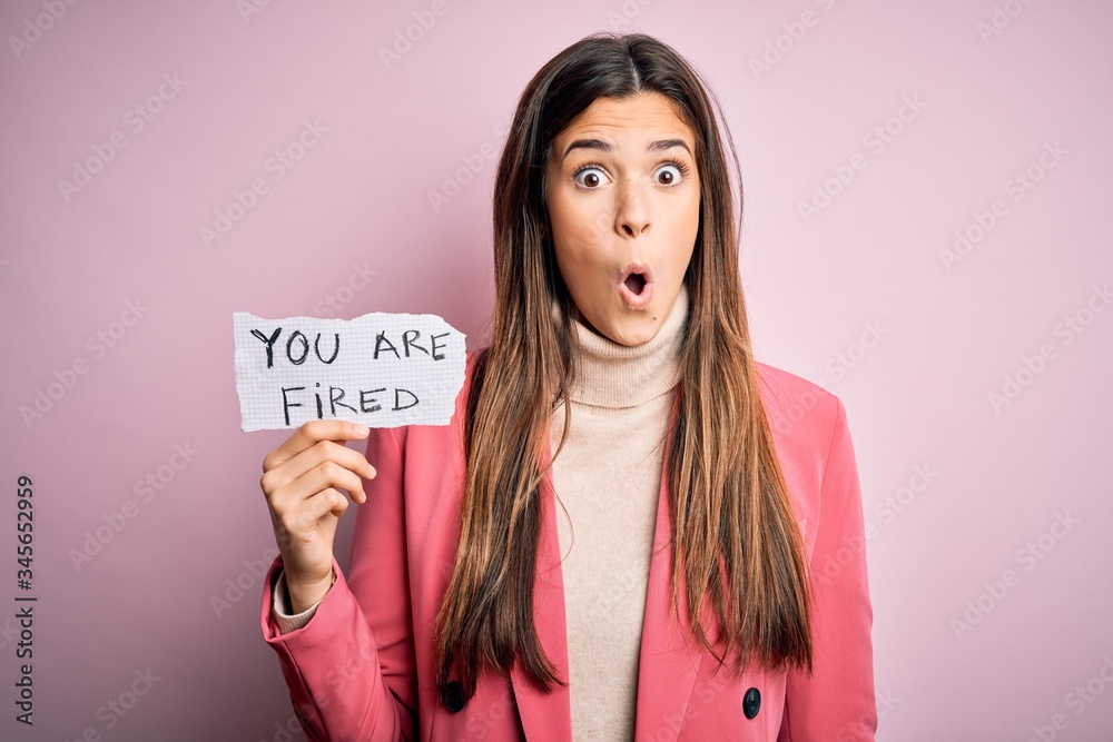Young beautiful girl holding paper with you are fired message over isolated pink background scared in shock with a surprise face, afraid and excited with fear expression Young beautiful girl holding paper with you are fired message over isolated pink background scared in shock with a surprise face, afraid and excited with fear expression