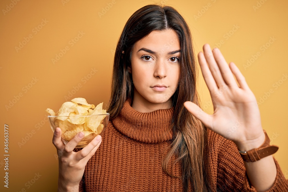 Young beautiful girl holding bowl with chips potatoes standing over yellow background with open hand doing stop sign with serious and confident expression, defense gesture Young beautiful girl holding bowl with chips potatoes standing over yellow background with open hand doing stop sign with serious and confident expression, defense gesture