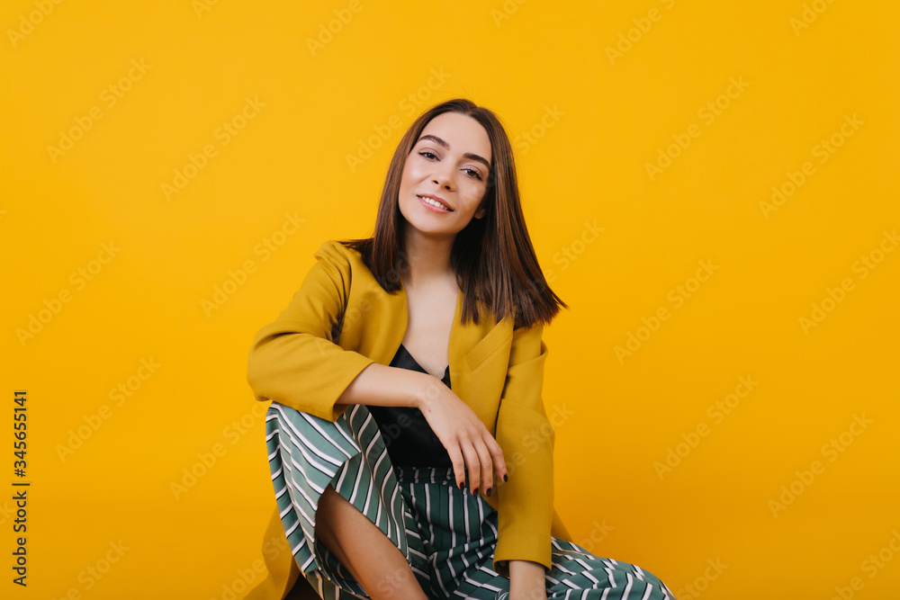 Enchanting girl in trendy jacket sitting on the floor in studio. Romantic young lady with brown hair posing with interedted smile. Enchanting girl in trendy jacket sitting on the floor in studio. Romantic young lady with brown hair posing with interedted smile.