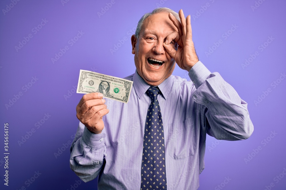 Senior grey haired man holding one dollar bank note over purple background with happy face smiling doing ok sign with hand on eye looking through fingers Senior grey haired man holding one dollar bank note over purple background with happy face smiling doing ok sign with hand on eye looking through fingers