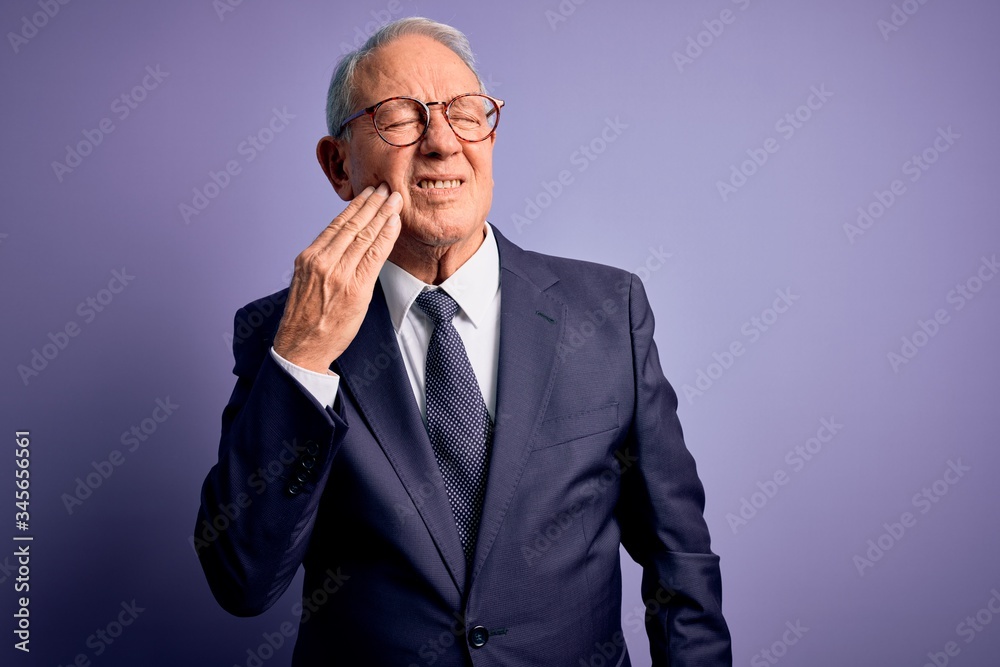 Grey haired senior business man wearing glasses and elegant suit and tie over purple background touching mouth with hand with painful expression because of toothache or dental illness on teeth. Grey haired senior business man wearing glasses and elegant suit and tie over purple background touching mouth with hand with painful expression because of toothache or dental illness on teeth.