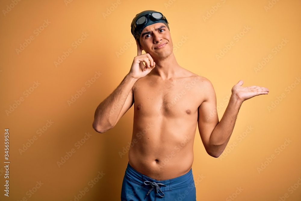 Young handsome man shirtless wearing swimsuit and swim cap over isolated yellow background confused and annoyed with open palm showing copy space and pointing finger to forehead. Think about it.