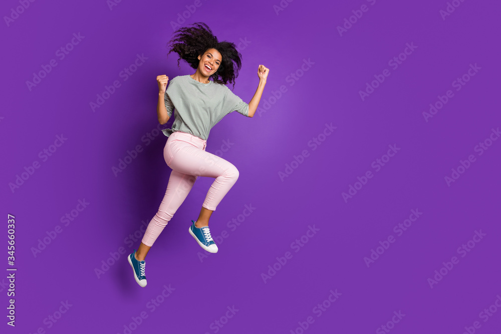 Full length profile side photo of cheerful funky afro american girl jump enjoy discounts win lottery raise fists scream yes wear striped shirt sneakers isolated violet color background