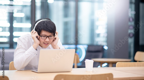 Young Asian man wearing headphones listening to music while working with laptop computer at home office. Relaxing at work concept