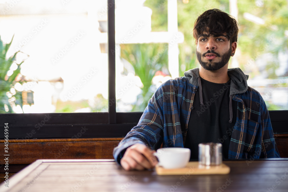 Young handsome bearded Indian hipster man thinking at the coffee shop Young handsome bearded Indian hipster man thinking at the coffee shop