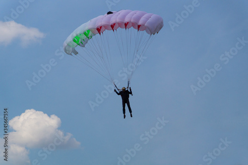 A skydiver in black outfit and black glasses flies across the sky with clouds on a white-red-green parachute