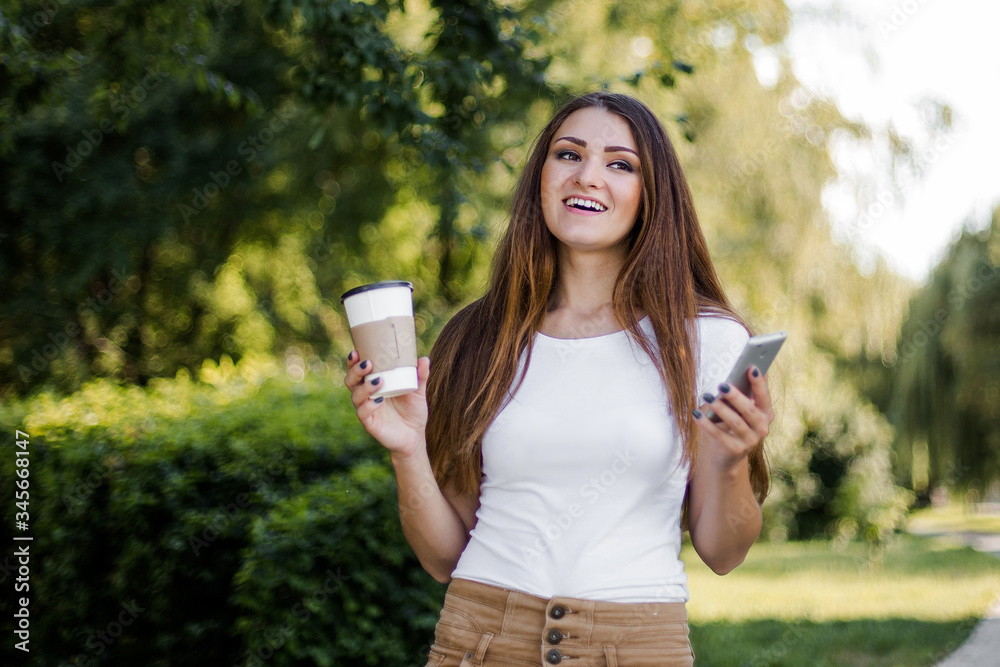 Young pretty woman using phone and drink coffee walking in summer park. Young pretty woman using phone and drink coffee walking in summer park.