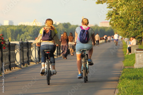 A cyclists woman pair rides together on bicycles along the embankment in Park on a summer day, back view on alleyway road, urban outdoor sports recreation
