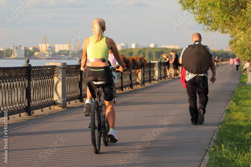A slender European sexy blonde young woman in sportswear rides a bicycle along the embankment on a Sunny summer evening, back view, urban outdoor active recreation