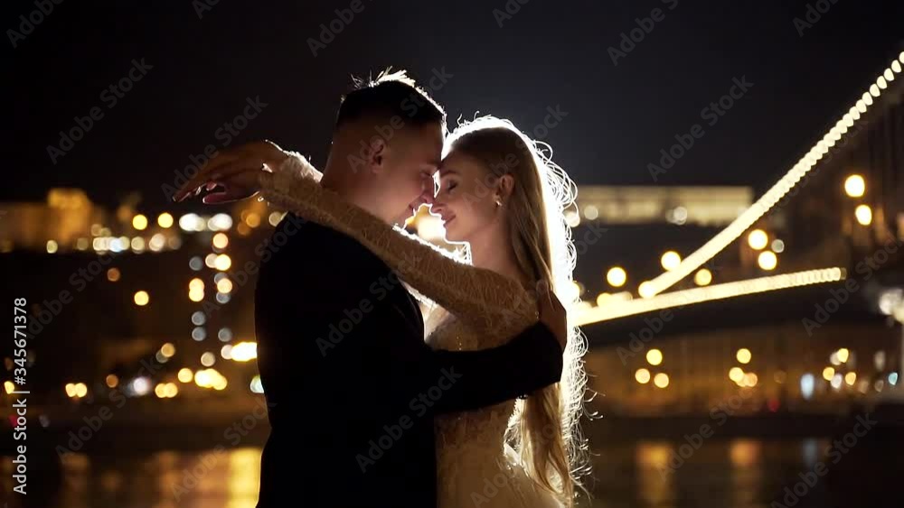 Portrait. wedding couple, bride and groom in white dress hugging, against the background of night old town, river and big bridge. bokeh lights. light. Evening. Slow motion. 
fashion, luxury lovers