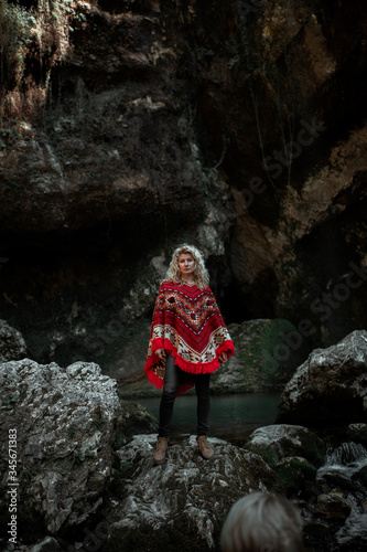 Attractive blonde curly young woman wearing red authentic poncho with ethnic pattern, in the beautiful stony gorge or cave 