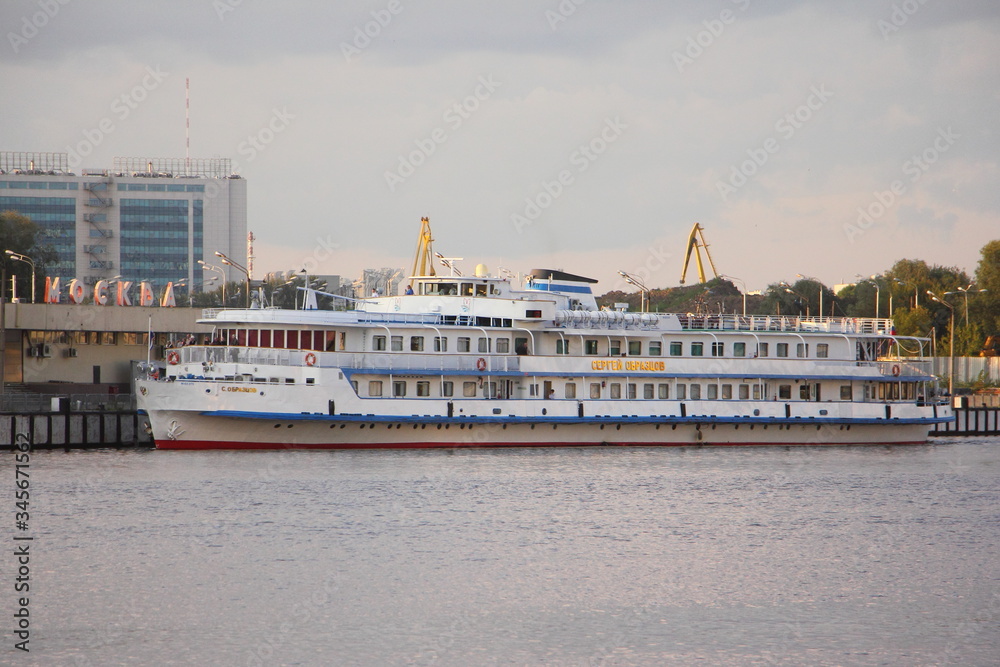 Moscow / Russia – 07 21 2019: White tourist ship Sergey Obraztsov near Pier South Port on Moscow River water and Embankment background on cloudy Summer day