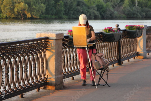 Young girl street artist draws on an easel on an empty alley in a Park on the waterfront on a Sunny summer evening, Amateur art outdoor photo