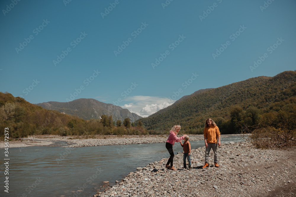 Fototapeta premium Beautiful young family, dad, mother and little son, having fun on the beach of mountain river near green summer forest with great view in the bright sunny day, blue sky backward