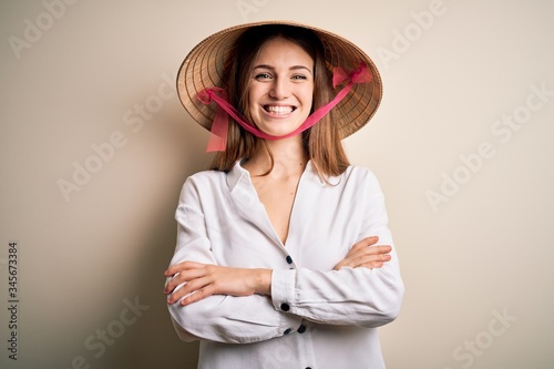 Young beautiful redhead woman wearing asian traditional conical hat over white background happy face smiling with crossed arms looking at the camera. Positive person.