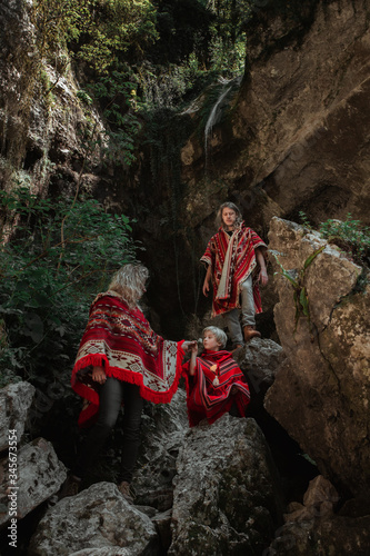 Beautiful happy caucasian family wearing authentic red poncho with ethnic pattern in the wild stony cave or gorge, dark stones backward