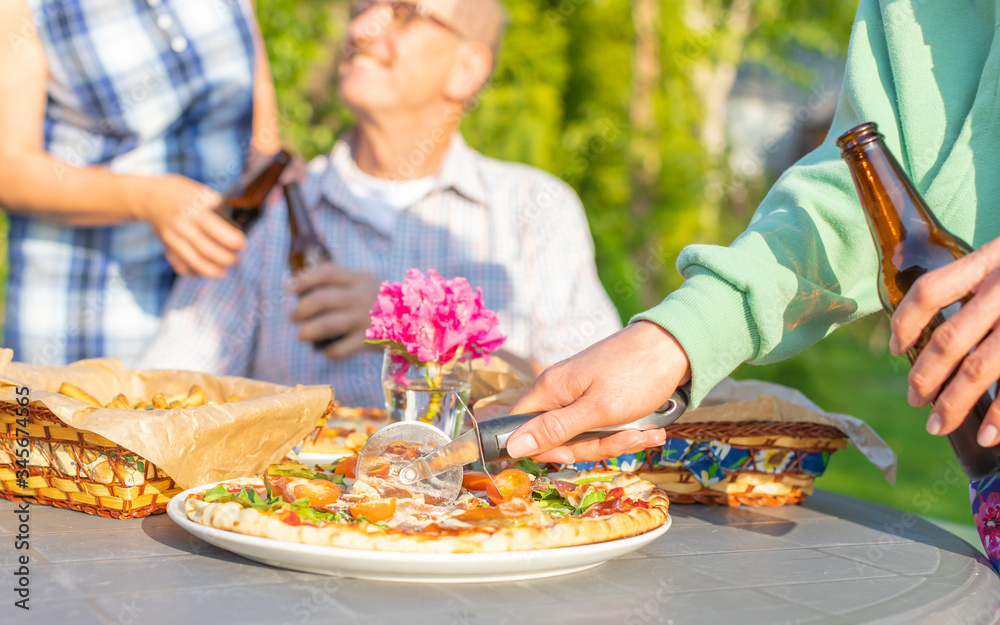 Cropped image of woman cutting pizza at family party while senior couple toasting with beer on background