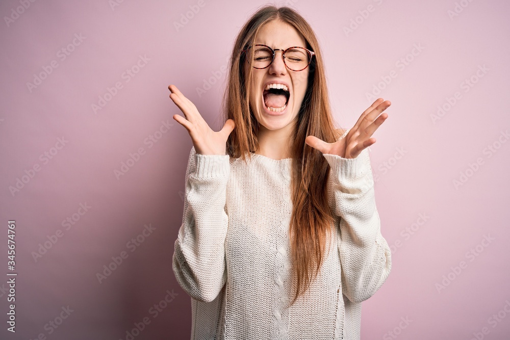 Young beautiful redhead woman wearing casual sweater and glasses over pink background celebrating mad and crazy for success with arms raised and closed eyes screaming excited. Winner concept Young beautiful redhead woman wearing casual sweater and glasses over pink background celebrating mad and crazy for success with arms raised and closed eyes screaming excited. Winner concept