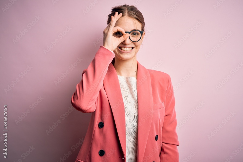 Young beautiful redhead woman wearing jacket and glasses over isolated pink background doing ok gesture with hand smiling, eye looking through fingers with happy face.