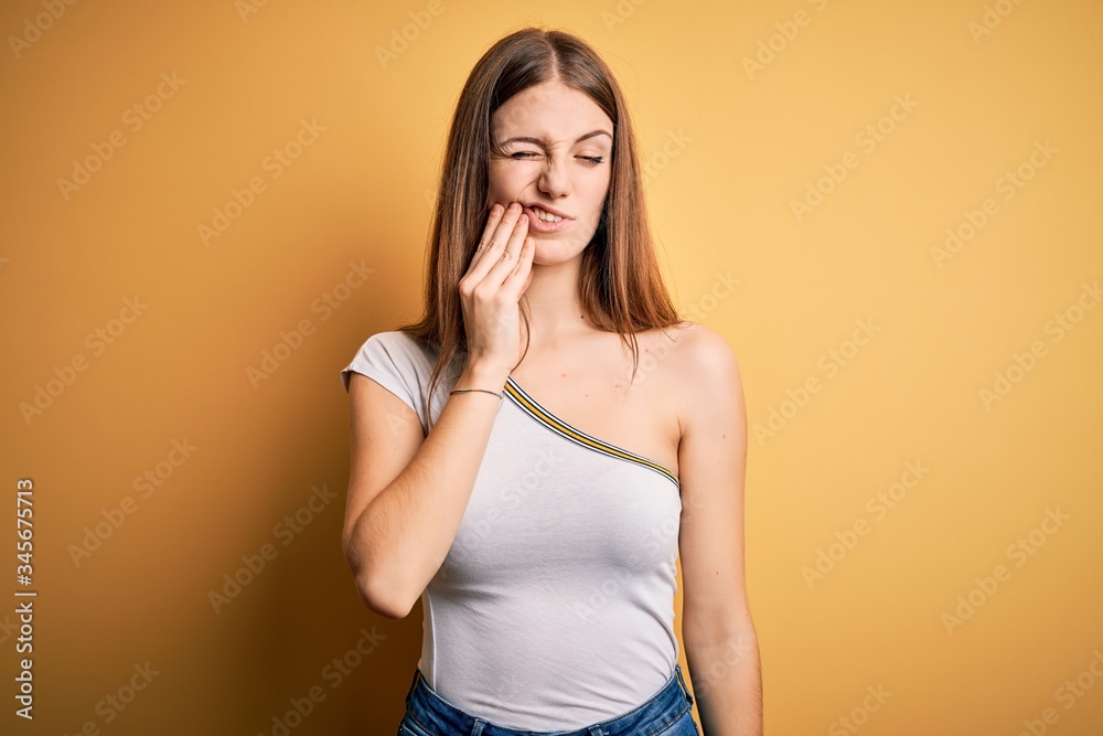 Young beautiful redhead woman wearing casual t-shirt over isolated yellow background touching mouth with hand with painful expression because of toothache or dental illness on teeth. Dentist