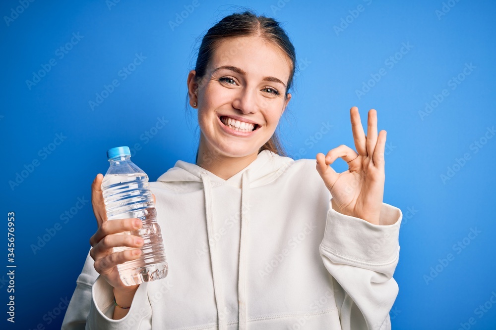 Young beautiful redhead woman doing sport drinking bottle of water over blue background doing ok sign with fingers, excellent symbol Young beautiful redhead woman doing sport drinking bottle of water over blue background doing ok sign with fingers, excellent symbol