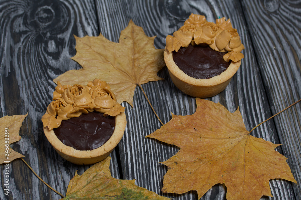 Tartlets with chocolate ganache. On autumn maple leaves. Decorated with oil cream flowers. The cream has a caramel color. On brushed pine boards painted black.