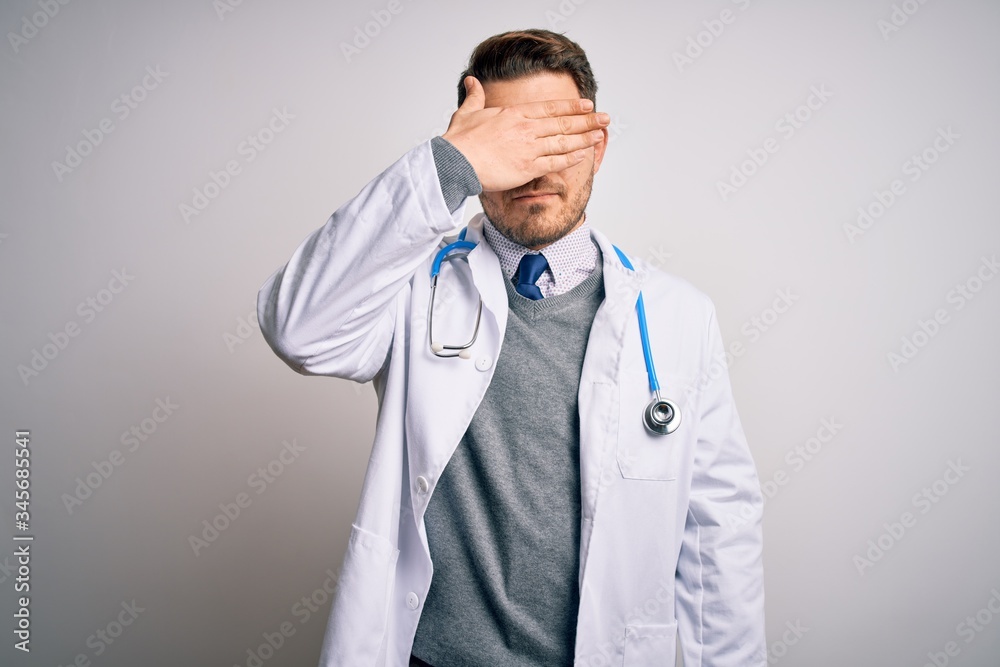 Young doctor man with blue eyes wearing medical coat and stethoscope over isolated background covering eyes with hand, looking serious and sad. Sightless, hiding and rejection concept Young doctor man with blue eyes wearing medical coat and stethoscope over isolated background covering eyes with hand, looking serious and sad. Sightless, hiding and rejection concept