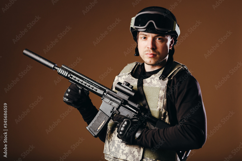 Soldier with gun is standing and looking on camera on brown background. Concept of war. Veterans, comrades, soldiers. Man in uniform. Soldier with gun is standing and looking on camera on brown background. Concept of war. Veterans, comrades, soldiers. Man in uniform.