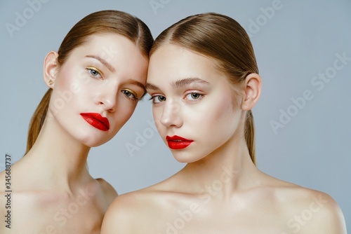 Close-up portrait of two gorgeous young fashion models with red lips posing grey background. Beautiful healthy face of the young pretty woman with fresh skin.