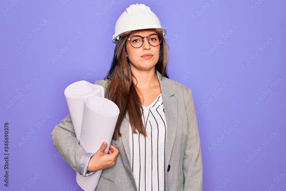 Young hispanic architect woman wearing builder hat and holding blueprints over yellow background with a confident expression on smart face thinking serious Young hispanic architect woman wearing builder hat and holding blueprints over yellow background with a confident expression on smart face thinking serious
