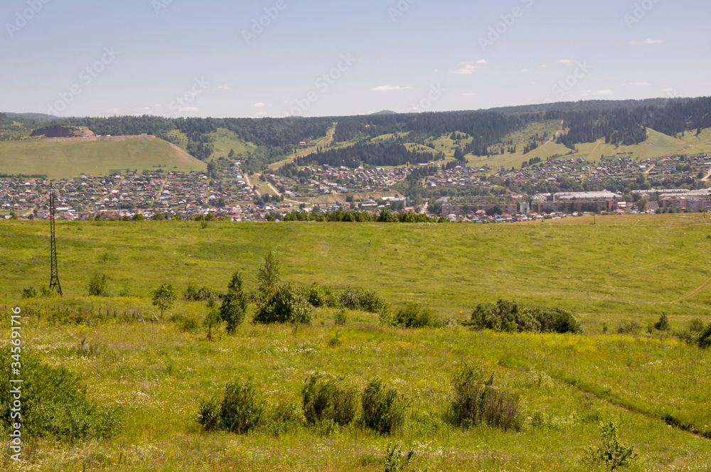 Suburb and villages. Big mountains and dark green forests far away. Trees and their shadows on the grass. Summer day with bright blue sky and huge white clouds on the landscape