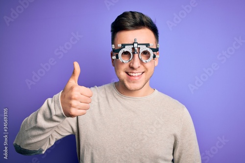 Young handsome caucasian man wearing optometrical glasses over purple background doing happy thumbs up gesture with hand. Approving expression looking at the camera showing success.