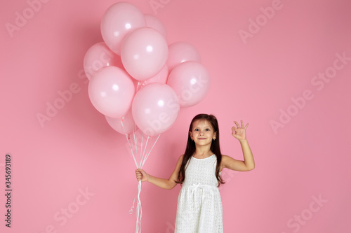 Smiling adorable little child girl with pastel pink air balloons showing Ok gesture isolated over pink background. Beautiful happy kid on a birthday party. space for text