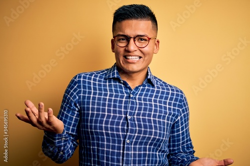 Young handsome latin man wearing casual shirt and glasses over yellow background smiling cheerful with open arms as friendly welcome, positive and confident greetings