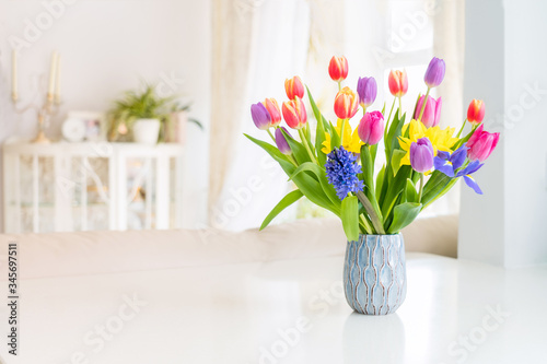 Fotografie Fresh spring colorful bouquet of tulips, daffodils, irises in vase standing on white marble table with light classic design living room background