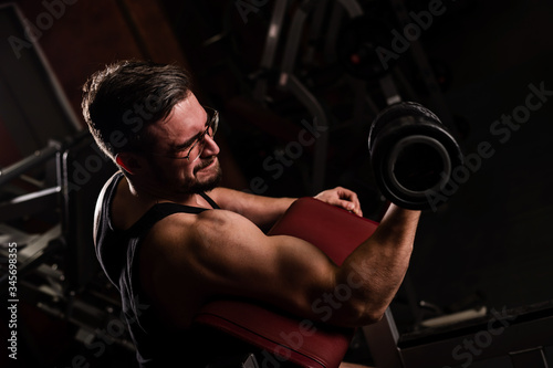 A handsome man with glasses doing an exercise for biceps with a barbell. The guy is engaged in bodybuilding. Trainer in the gym with muscular arms.
