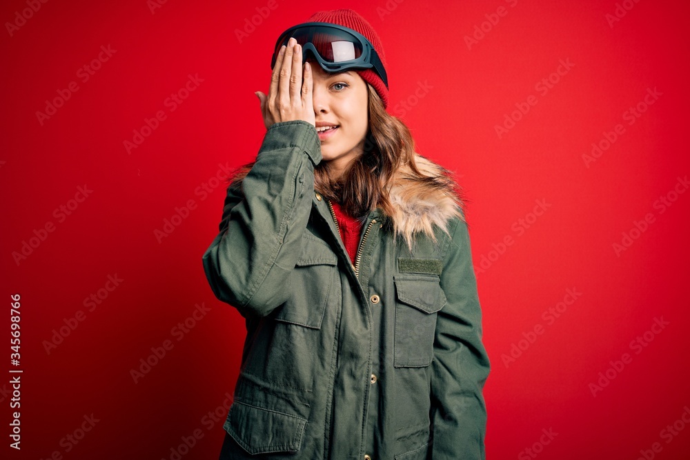 Young blonde girl wearing ski glasses and winter coat for ski weather over red background covering one eye with hand, confident smile on face and surprise emotion. Young blonde girl wearing ski glasses and winter coat for ski weather over red background covering one eye with hand, confident smile on face and surprise emotion.