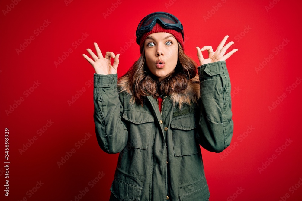 Young blonde girl wearing ski glasses and winter coat for ski weather over red background looking surprised and shocked doing ok approval symbol with fingers. Crazy expression Young blonde girl wearing ski glasses and winter coat for ski weather over red background looking surprised and shocked doing ok approval symbol with fingers. Crazy expression