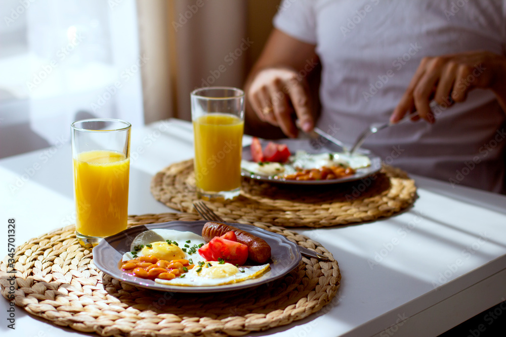 Man eats scrambled eggs for breakfast in the kitchen. Breakfast on plates of fried eggs, beans, sausages, tomatoes, orange juice in a glass on a table.