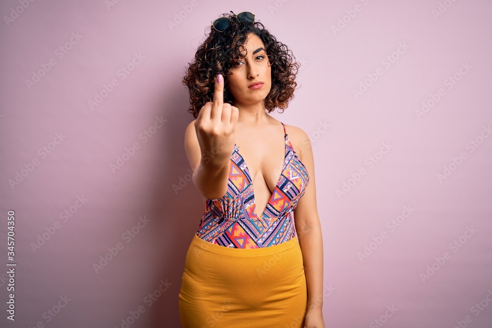 Young beautiful arab woman on vacation wearing swimsuit and sunglasses over pink background Showing middle finger, impolite and rude fuck off expression Young beautiful arab woman on vacation wearing swimsuit and sunglasses over pink background Showing middle finger, impolite and rude fuck off expression